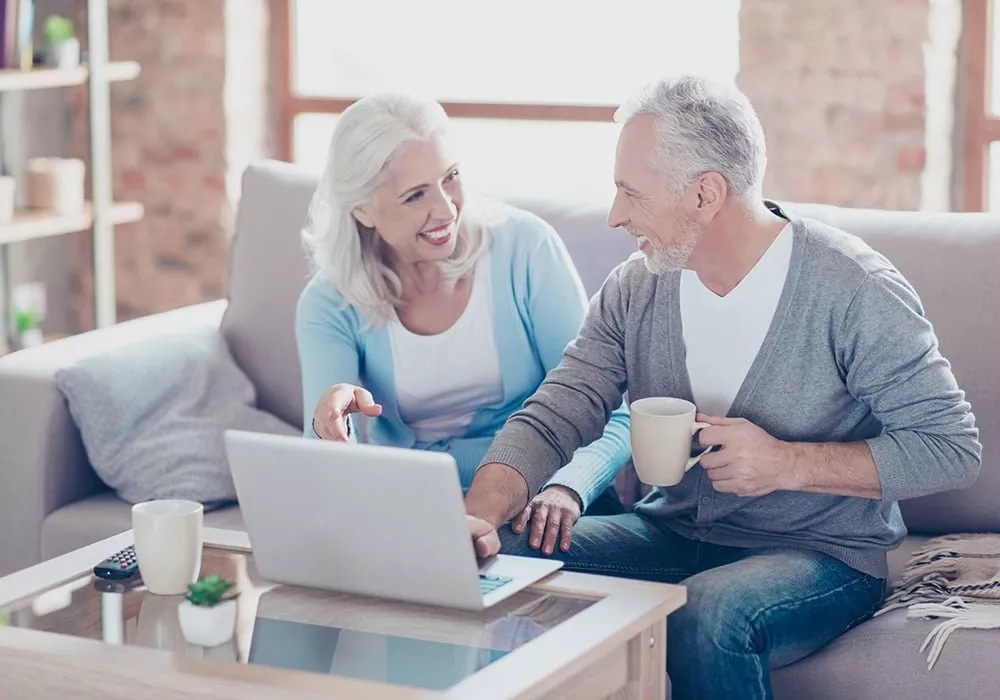 Two older adults sit on a sofa, smiling whilst looking at a laptop. One holds a mug, and both appear engaged in a conversation as they explore care information together.